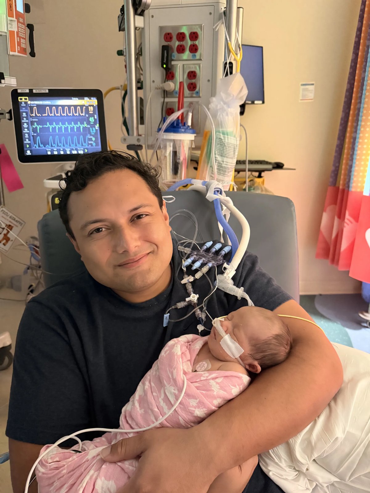 Tala's dad smiling as he holds Tala in his arms, Tala swaddled in pink with her breathing tube, monitors behind them