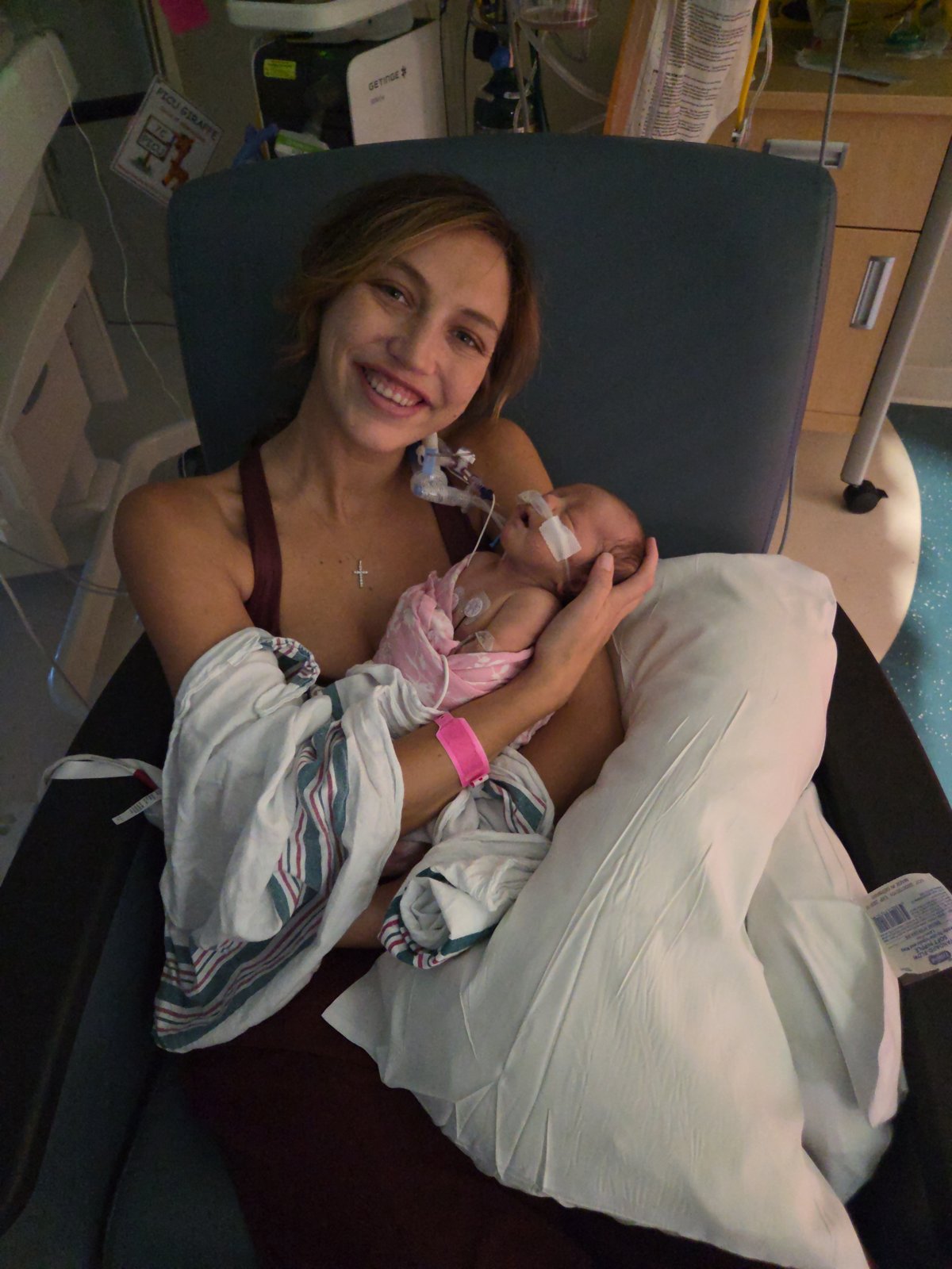 Tala's mom smiling as she holds Tala against her chest in a hospital chair, Tala swaddled in pink with her breathing tube